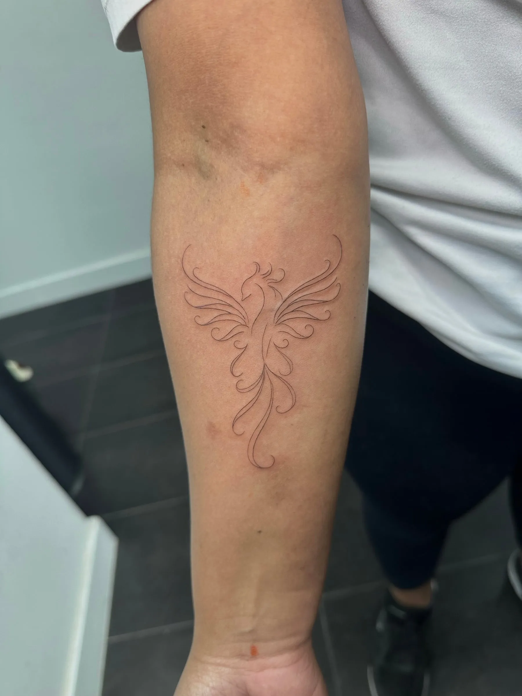 Ave fénix con las alas abiertas en línea fina y trazos ornamentales, en el antebrazo